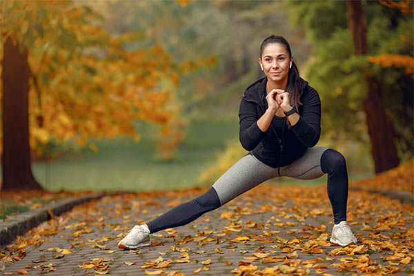 A woman doing a side lunge stretch on a leaf-covered path in a park during autumn, wearing gym cloths suitable for outdoor workouts.