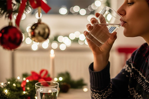 A woman drinking a glass of water beside a beautifully decorated Christmas tree with lights and ornaments. Rest During the Festive Season